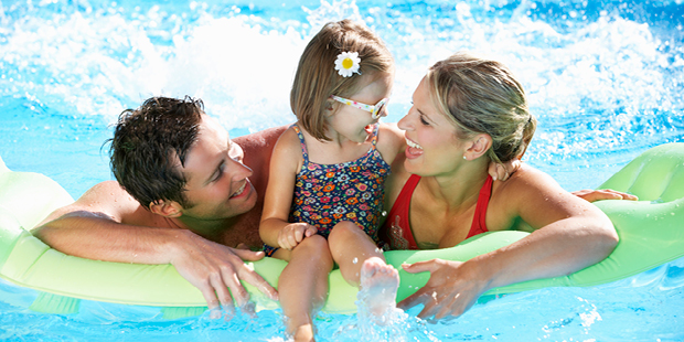 Bilden visar två föräldrar och ett barn som sitter på en badmadrass i poolen