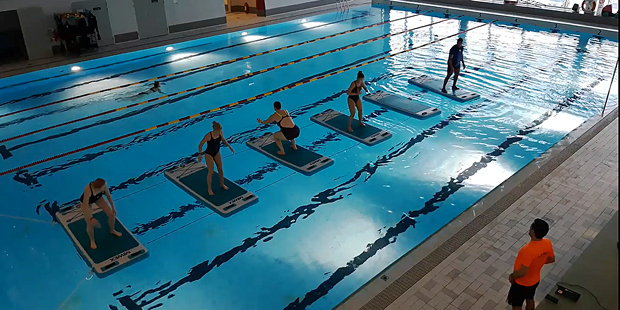 Bilden visar en pool där några personer står på varsin träningsmatta i vattnet framför en instruktör